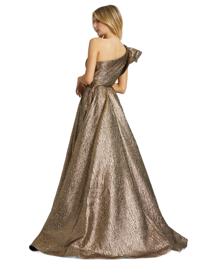 A woman wears the Mac Duggal 67297 Metallic Long Prom Ball Gown, featuring a fitted bodice and full floor-length skirt. She faces away as the shimmering gold textured evening dress catches the light.